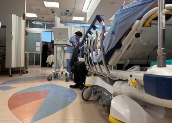 A stretcher in a hospital emergency department hallway with someone wearing scrubs in the distance.