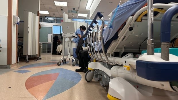 A stretcher in a hospital emergency department hallway with someone wearing scrubs in the distance.