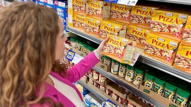 A woman with long curly hair and a pink blazer inspects a package of pizza dough on a wall at the grocery store.
