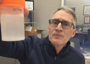 A man with gray hair and glasses wearing a blue sweater holds up a cloudy bottle with white powder in liquid.