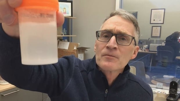A man with gray hair and glasses wearing a blue sweater holds up a cloudy bottle with white powder in liquid.