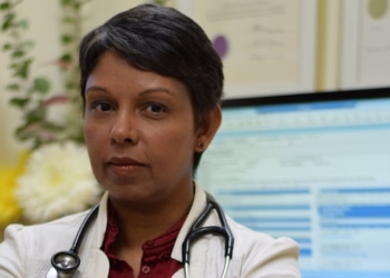 A short-haired woman in a lab coat with a stehoscope around her neck sits facing away from a computer
