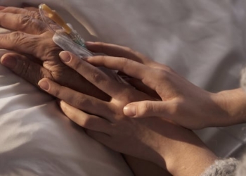 The hands of an adult hold the hand of an older person, who is hooked up to intravenous and lying in a bed.