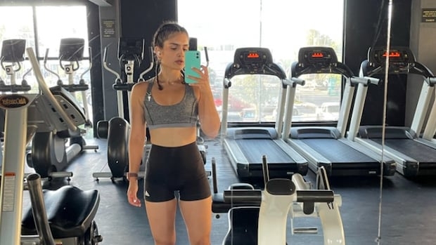 A fit woman in form-fitting shorts and a crop top takes a full-body selfie of herself in the mirrors that also show fitness machines behind her.