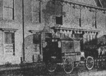 A grainy black-and-white photo shows an early, horse-drawn ambulance parked on a street in front of a brick building. The word