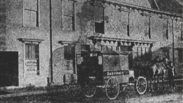 A grainy black-and-white photo shows an early, horse-drawn ambulance parked on a street in front of a brick building. The word
