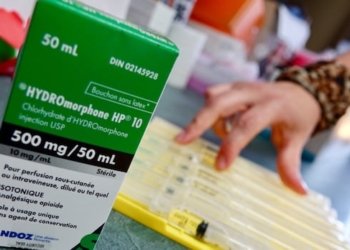 A green box of medication is pictured. A person's hand is visible in the background with  a tray of medical supplies.