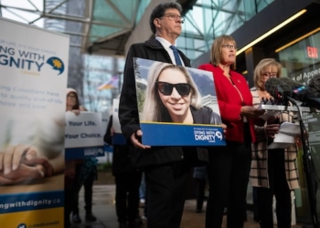 A group of people stand in front of a banner marked 'Dying with Dignity', holding a photo of a blond woman.