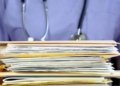 A stack of medical files are held by someone wearing blue scrubs and a stethoscope.