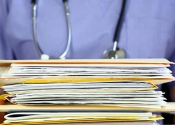 A stack of medical files are held by someone wearing blue scrubs and a stethoscope.