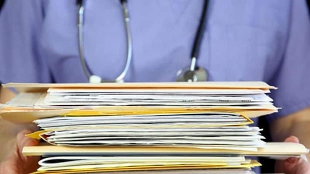 A stack of medical files are held by someone wearing blue scrubs and a stethoscope.