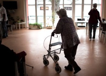 An older woman uses a walker in a room with a large window, dining tables to the right and chairs to the left.