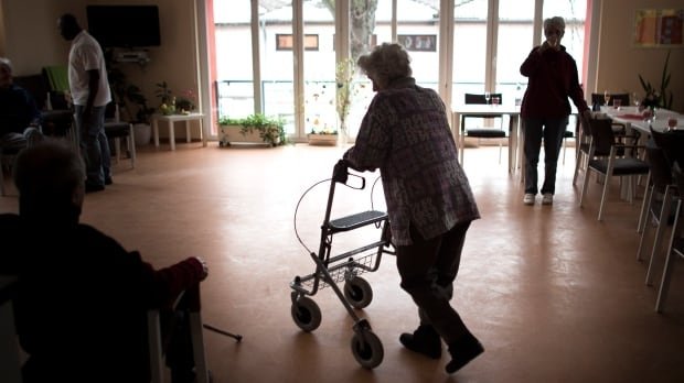An older woman uses a walker in a room with a large window, dining tables to the right and chairs to the left.