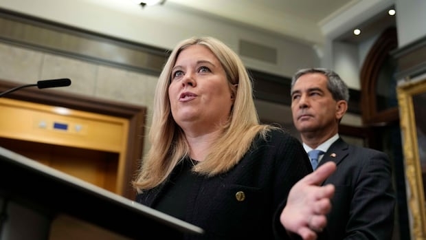 A woman speaks at a lectern while a man stands behind her.