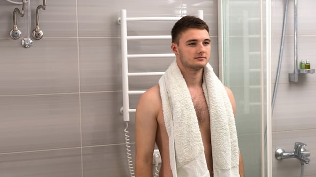 A young man with a towel around his shoulders stands in a bathroom.