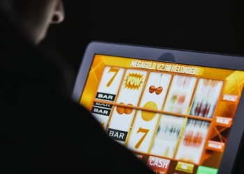 The back of a person's shoulder in a dark room, hunched over a screen with a virtual slot machine spinning