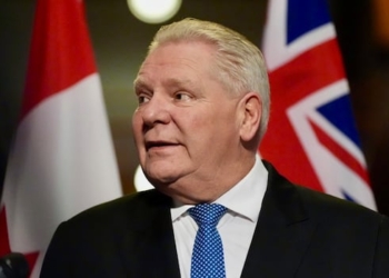A man standing in front of a row of flags looks to his right as a reporter asks him a question.