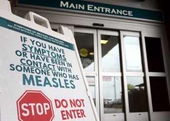 A white sign in front of a hospital entrance tells people not to enter if they have symptoms or have been in contact with someone who has measles.