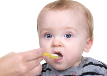 A baby with blue eyes being fed rice cereal with a yellow spoon.