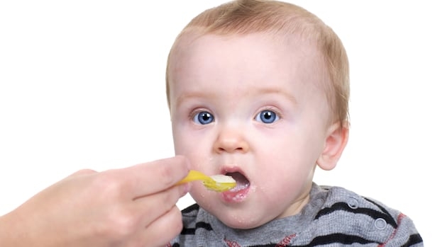 A baby with blue eyes being fed rice cereal with a yellow spoon.