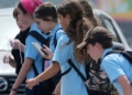 A group of kids in light blue uniform polo shifts cross a street, with one girl looking down at a cellphone while doing so.