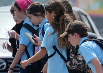 A group of kids in light blue uniform polo shifts cross a street, with one girl looking down at a cellphone while doing so.