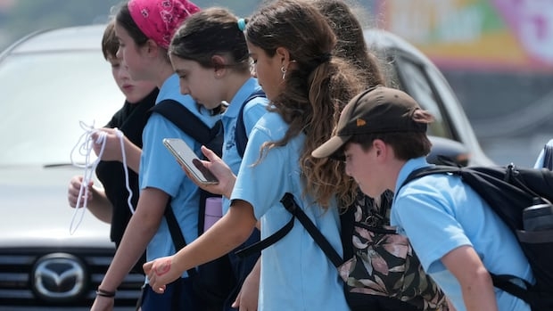 A group of kids in light blue uniform polo shifts cross a street, with one girl looking down at a cellphone while doing so.