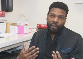 A man in his thirties sits in a medical office speaking with a reporter