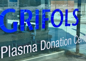 A sign saying Grifols appears on the glass exterior wall of a building with snow seen on the ground near the building.