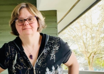 A woman stands on a porch outside, posing for a photo. She is in her 40s and has Down syndrome