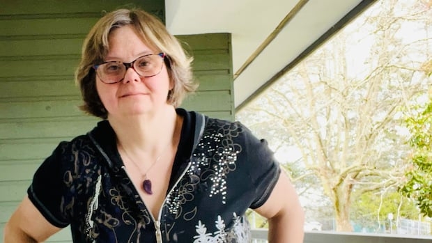 A woman stands on a porch outside, posing for a photo. She is in her 40s and has Down syndrome