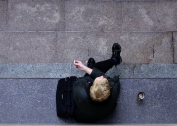 A person dressed all in black is shown from above, sitting on a stone bench smoking a cigarette. A black bag is beside them, along with a drink.
