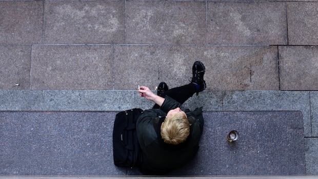 A person dressed all in black is shown from above, sitting on a stone bench smoking a cigarette. A black bag is beside them, along with a drink.