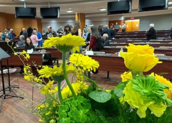 A lecture hall with wooden desks. A vase of yellow flowers with greenery sits in the foreground.
