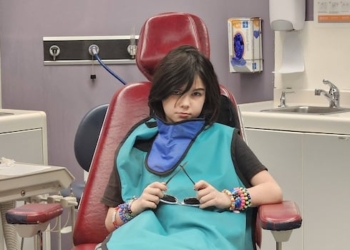 A child with shoulder length brown hair sits in a dentist chair.