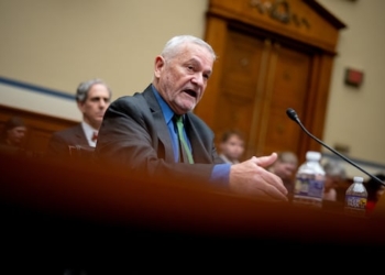 A man in a black suit and blue shirt speaks at a hearing.