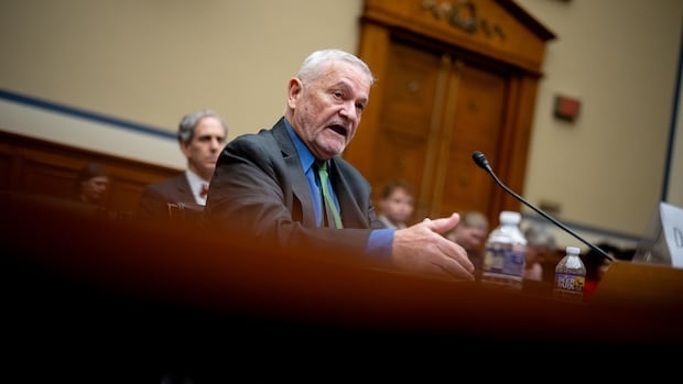 A man in a black suit and blue shirt speaks at a hearing.