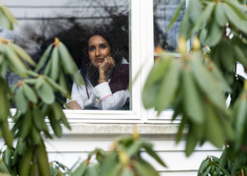 A woman looks out her window. Blurred greenery is seen in the foreground.