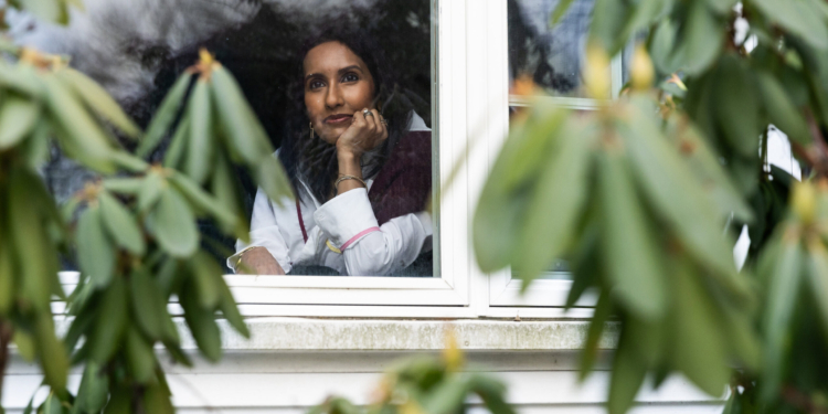 A woman looks out her window. Blurred greenery is seen in the foreground.