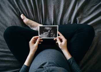 A high-angle photograph of a pregnant person sitting cross-legged on a bed and holds a sonogram of a baby.