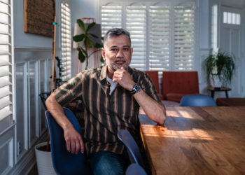 A South Asian man sits by the window at a table. He rests his elbow on the table with his hand on his chin. He looks contemplative.