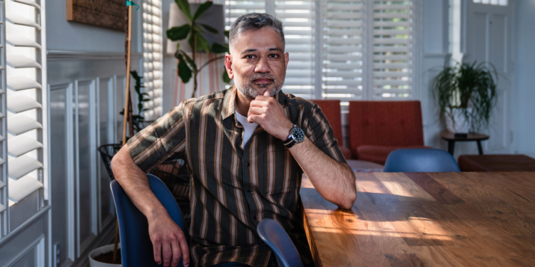 A South Asian man sits by the window at a table. He rests his elbow on the table with his hand on his chin. He looks contemplative.