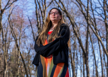 A woman stands outside in a wooded area.