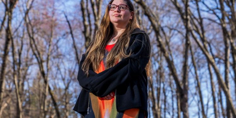 A woman stands outside in a wooded area.