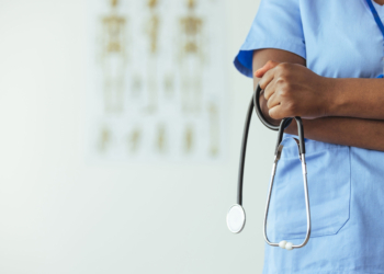 An unidentifiable medical professional stands to the side of frame in blue scrubs and holds a stethoscope.