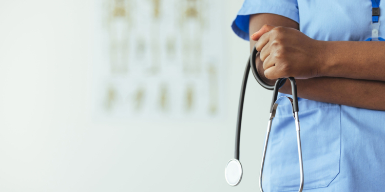 An unidentifiable medical professional stands to the side of frame in blue scrubs and holds a stethoscope.