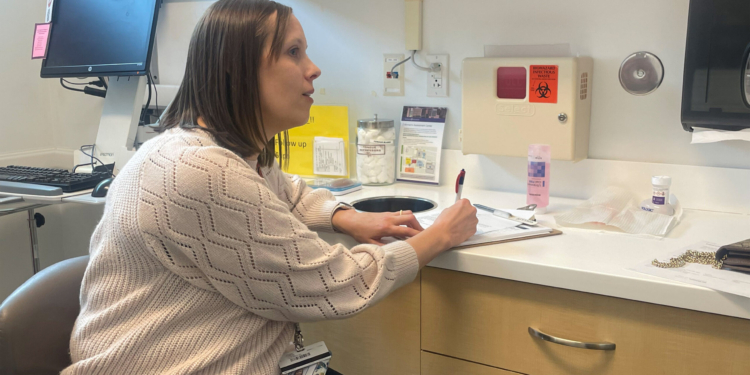 A doctor writes notes in an examination room while talking to a patient out of frame.