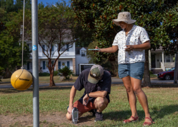 Two adults work outside. One on the left uses a device pressed to the ground to test for lead. Another holds out her arm, phone in hand, to get coordinates.