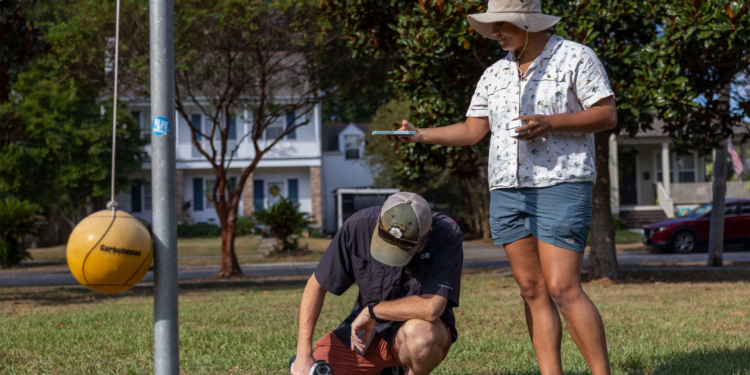 Two adults work outside. One on the left uses a device pressed to the ground to test for lead. Another holds out her arm, phone in hand, to get coordinates.