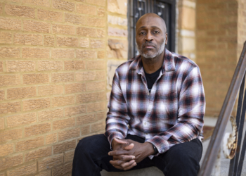 A man wearing a plaid button-up shirt sits on the front step of a home for a portrait